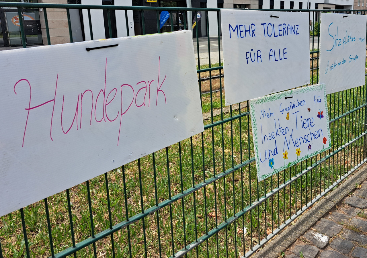 Zaun mit handgemalten Plakaten und Wünschen für die Innenstadt von morgen. Darauf steht "Hundepark", "Mehr Toleranz für alle", "Mehr Grünflächen für Insekten, Tiere und Menschen", "Sitzplätze" - die Plakate sind bei einem Aktionstag zur Innenstadtgestaltung in Griesheim entstanden.