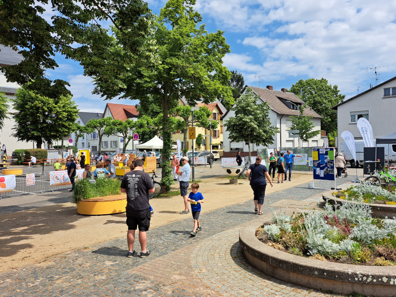 Bild der Auftaktveranstaltung zum Projekt "Griesheim. Innen Drin". Kinder und Erwachsene auf einem Platz mit Bäumen, Stadtmöbeln, Infotafeln.