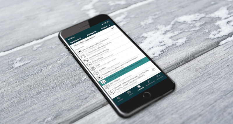 Smartphone auf Holz, auf dem Display sind die verschiedenen Meldekategorien vom Mängelmelder der EGLV zu sehen.